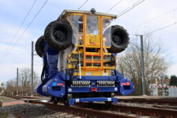 Le MASS de Novium, machine rail-route bleue et jaune, positionné sur une voie ferrée pour la stabilisation du ballast.