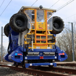 Le MASS de Novium, machine rail-route bleue et jaune, positionné sur une voie ferrée pour la stabilisation du ballast.